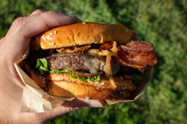 Bir el taze pişmiş, sulu bir batı barbekü burgeri, peynirli, kızarmış, soğanlı, marullu, pastırmalı, barbekü soslu yeşil çimenli bir arka planda susamlı çöreğin üstünde..