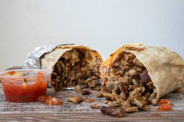 Folyoya sarılmış karne asada bifteği dürümü. İkiye bölünmüş ve içinden et dökülüyor. Masada patates kızartması ve salsa sosu var..