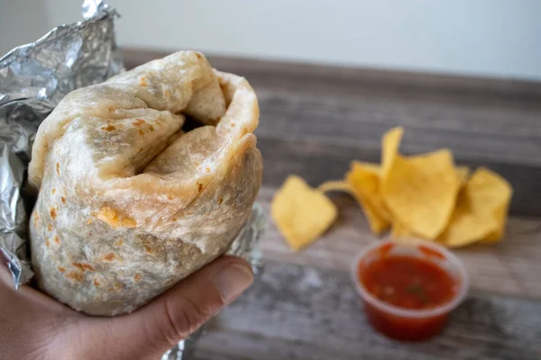 Kaliforniya usulü bir burritoyu tahta bir masanın üstünde patates kızartması ve salsa sosuyla sarmalayan bir el.