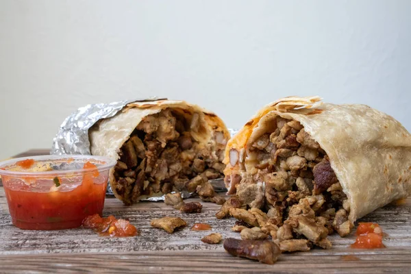 Folyoya sarılmış karne asada bifteği dürümü. İkiye bölünmüş ve içinden et dökülüyor. Masada patates kızartması ve salsa sosu var..