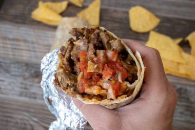Izgara biftekli burrito ve salsa soslu tortilla cipsi tutan bir el.