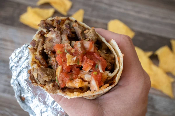 Izgara biftekli burrito ve salsa soslu tortilla cipsi tutan bir el.
