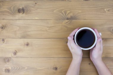 Kadınlar elleri süveter içinde, siyah ahşap masada bir fincan kahve tutuyorlar.