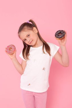 Lezzetli çörekler tutan küçük güzel bir kız başparmağıyla yan tarafa bakıyor ve ağzı açık gülümsüyor.