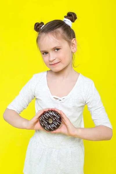 Uma menina bonita de aparência europeia em um vestido azul com listras brancas morde um donut em esmalte brilhante. Criança alegre alegre alegre alegre e alegre com um rosto esfarrapado em um fundo laranja — Fotografia de Stock