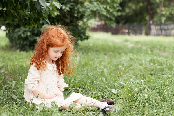 A smiling red-haired little girl with loose curly long hair in a dress walks in a city park and looks at the green leaves of a tree — Foto de Stock