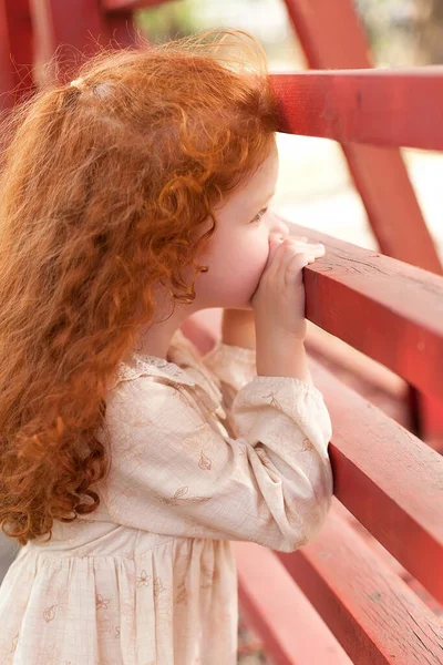 A beautiful little girl with red curly hair in a light dress, walking outdoors in a city park, looking at the sky. Portrait of a small child. — Foto de Stock