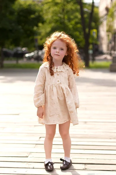 Portrait of a cute red-haired little girl holding hands near her face and dreaming, looking at something cute and cute. Shooting in a city park. — Foto de Stock