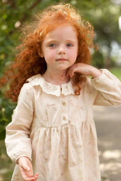 A little girl with red curly hair in a dress poses among wild flowers in a field at sunset. Portrait. Imagen de archivo