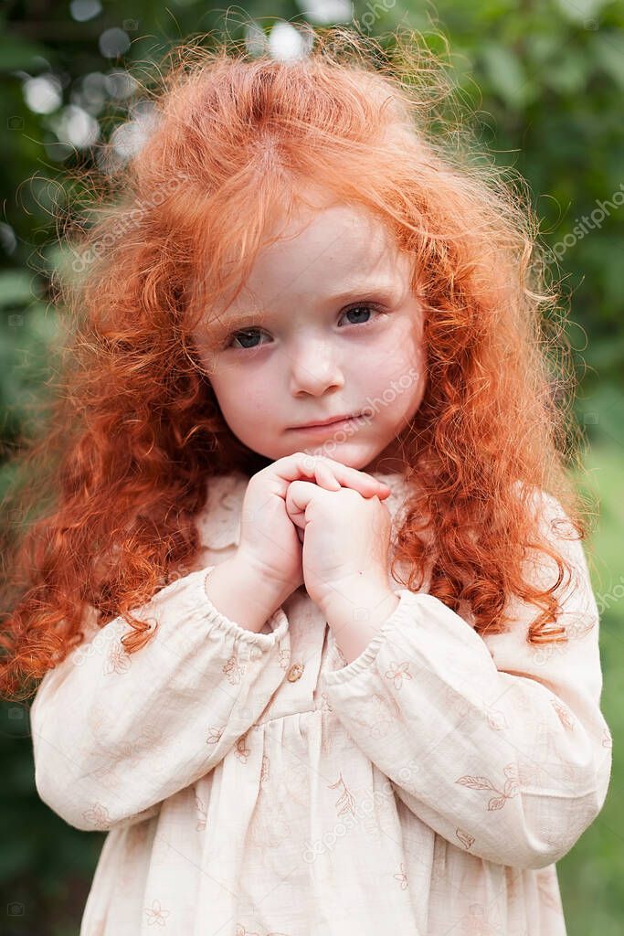 Portrait of a cute red-haired little girl holding hands near her face and dreaming, looking at something cute and cute. Shooting in a city park.