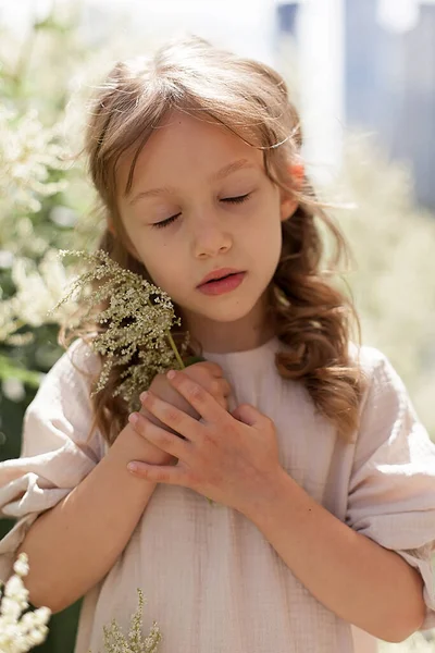 A romantic portrait of a little girl in a beige dress with a flower in her hands in a city park. The child has closed his eyes and is dreaming, holding a flower to his face. — Stock Photo, Image