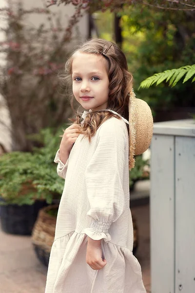 Забавна мила дівчина 5-6 років, в бежевій сукні і солом'яному капелюсі, позує в парку на тлі природи. Щастя. Літній сезон . Стокова Картинка