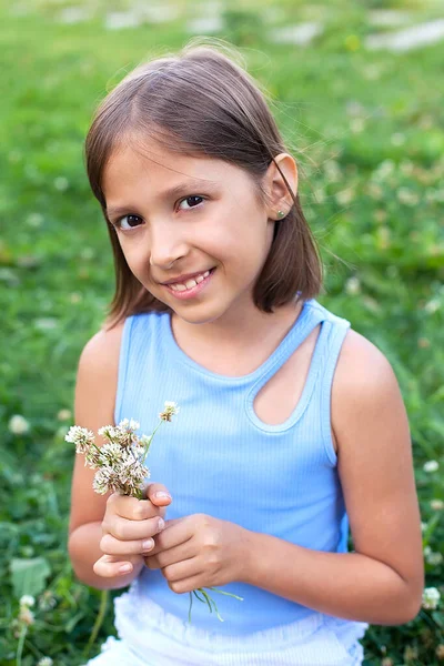Uma garotinha fofa em um prado em um dia de verão sorri e olha para a câmera, segurando um buquê de flores de verão em suas mãos. — Fotografia de Stock