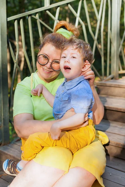 Porträt eines charmanten kleinen behinderten Jungen, der in den Armen seiner liebenden Mutter auf dem grünen Gras im Park liegt. Behinderung. Mütter lieben. Familienförderung. — Stockfoto
