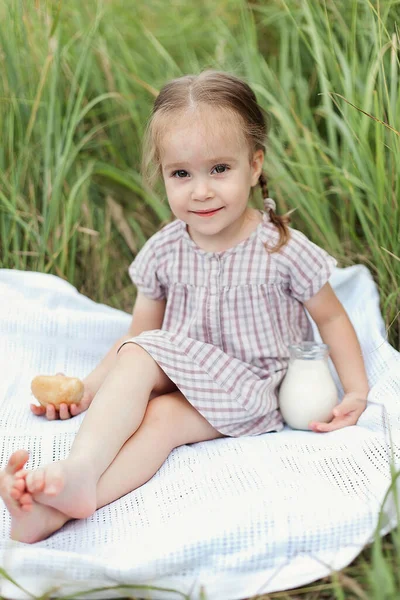 Um bebê segura um jarro de leite em suas mãos em um dia de verão em um campo verde. Uma menina está sentada em um cobertor e desfrutando da natureza — Fotografia de Stock