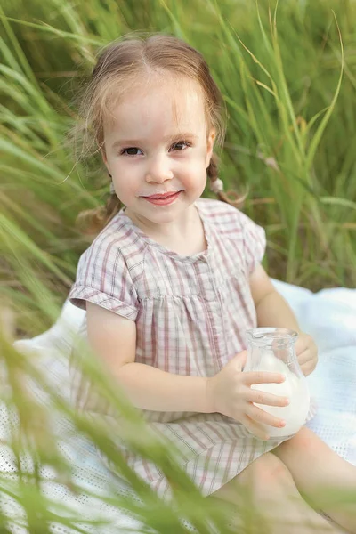 Ребенок держит в руках кувшин молока в летний день на зеленом поле. Маленькая девочка сидит на одеяле и наслаждается природой — стоковое фото