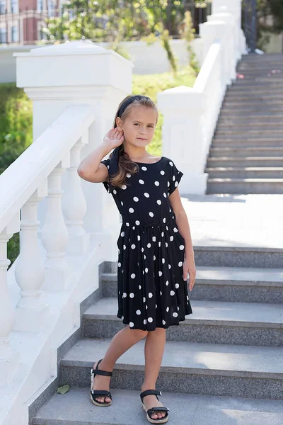 9歳の美しい少女が黒のドレスに身を包み ポルカの点が美しい白い階段に立って笑顔でカメラを見ています 夏の気分 — ストック写真