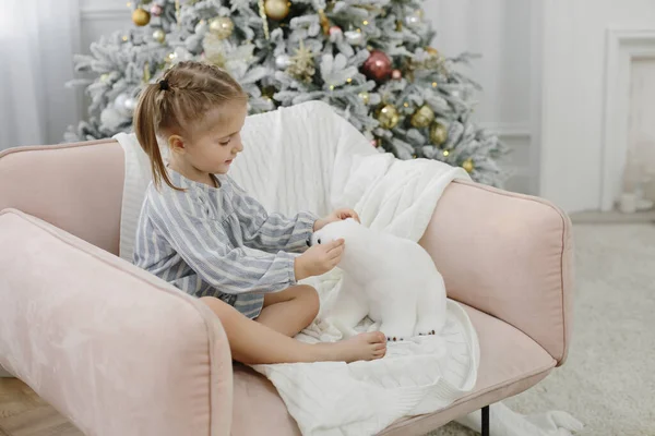 A three-year-old girl in a dress sits in a pink armchair on a New Year tree. A child plays with a polar bear in a light interior. Christmas and New Year concept.