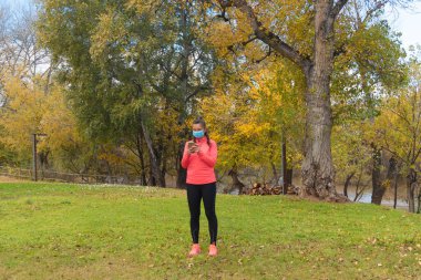 Sonbaharda bir parkın ortasında spor giyinmiş bir kadın kendini koronavirüsten korumak için ameliyat maskesi takarak cep telefonunu kontrol ediyor.