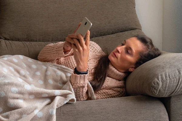 Woman lying on the sofa and covered with a blanket checks the mobile concentrated