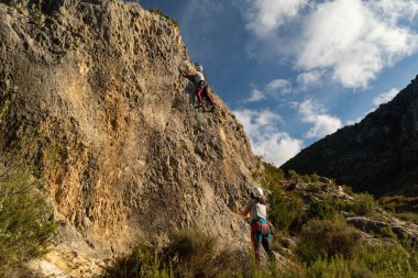 Dağcı çocuk tırmanma yolunun sonuna yaklaşırken bir kız onu aşağıdan bağlıyor.