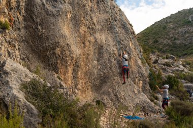 Genç kız, partneri onu bağlarken bir kaya duvarına tırmanmaya başlıyor.
