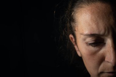 Çok loş aydınlatılmış bir kadının karanlık bir portresinin yakın çekimi. Kadın somurtuyor ve aşağıya bakıyor. Üzüntü ya da endişe duyuyor.