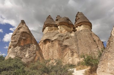 Dev mantarlara benzer keskin tepeleri olan inanılmaz yüksek uçurumlar bulutlu bir gökyüzünün arka planına karşı. Vadide yeşil çalılar yetişir. Kapadokya. Türkiye                               