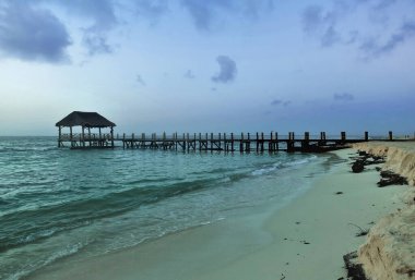 Turkuaz okyanusun üzerinde ahşap bir patika var ve bir de tepe örtüsü. Şeffaf dalgalar kumsalda yuvarlanıyor. Gökyüzünde ışık bulutları var. Meksika.