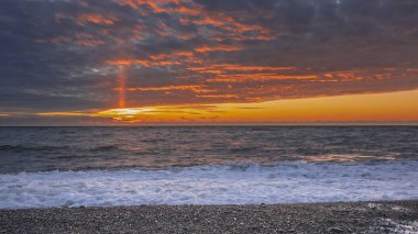 Deniz üzerinde parlak, dramatik bir gün batımı. Batan güneşin bir ışını mavi bulutları kırmızı ışıkla aydınlattı. Bir çakıl taşı plajında, beyaz bir dalga köpüğü. - Rusya. Soçi. Karadeniz