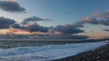 İyi akşamlar. Batan gökyüzünde, pembe ve mavi bulutlar. Denizde küçük dalgalar. Sörf, kumsaldaki çakıl taşlarında köpük bırakır. Uzakta pelerinin dış hatları. Karadeniz