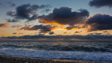 Akşam alacakaranlığı. Batan güneş mor bulutları turuncuyla aydınlatır. Deniz dalgaları kumsaldaki çakıl taşlarına beyaz köpük bırakır. Karadeniz.
