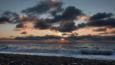 Gün batımı. Güneş ışınları koyu mavi bulutların arasından geçer ve onları turuncuyla aydınlatır. Deniz dalgaları köpükle yuvarlanır ve çakıl taşı plajına püskürtülür. Karadeniz.