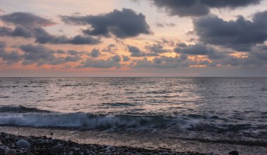 Denizin üzerinde gün batımı. Koyu mavi kümülüs bulutları pembe-turuncu gökyüzünde süzülür. Kumsaldaki çakıl taşlarında köpüren dalgalar. Karadeniz