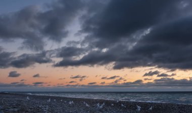 Pembeleşen günbatımı gökyüzünde, koyu mavi bulutlar. Denizde küçük dalgalar. Bir martı sürüsü kumsal çakıl taşlarında dinleniyor. Karadeniz