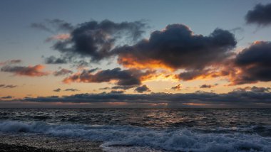 Karadeniz üzerinde yakıcı gün batımı. Mavi gökyüzündeki kara bulutlar turuncuyla işaretlenmiştir. Kumsalda köpüren dalgalar.