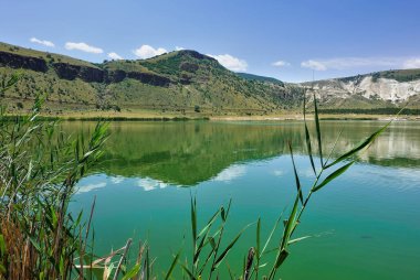The volcanic Lake Nar is surrounded by mountain slopes. Specular reflection in emerald water. In the foreground there is a reed. Clouds in the blue sky. Turkey. Cappadocia clipart