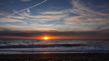 İyi akşamlar. Güneş ufukta batıyor. Mavi gökyüzü turuncu ile aydınlatılmış. Deniz dalgaları kıyıda köpürüyor. Su ve çakıl taşları üzerinde güneşli bir yol. Karadeniz.