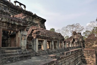 Angkor 'daki antik Baphuon tapınağı. Taş teraslar, boş pencereler, zamanla hasar görmüş. Duvarlar oymalar ve kabartmalarla süslenmiş. Arka plan açık mavi bir gökyüzü, ağaçlar. Kamboçya..