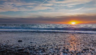 Açık bulutlu mavi günbatımı gökyüzü turuncuyla vurgulanıyor. Güneş batıyor. Turkuaz deniz dalgaları çakıl taşlarında köpük bırakır. Güneşli yol parlıyor. Karadeniz.