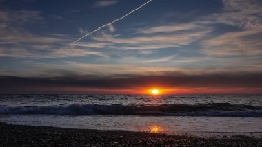 Denizin üzerinde parlak gün batımı. Mavi gökyüzünde bulutlar ve bir uçak yolu var. Güneş ufukta batıyor. Dalga çakıl taşı plajına yuvarlanıyor. Güneşli yol.