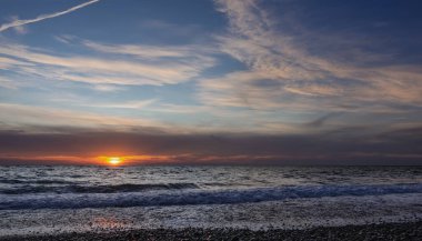 Deniz kenarında akşam alacakaranlığı. Mavi gökyüzünde sirus bulutları, uçak pisti. Ufkun üzerinde batan güneşten gelen turuncu bir ışık var. Dalgalar kumsaldaki çakıl taşlarında köpük bırakır. Karadeniz