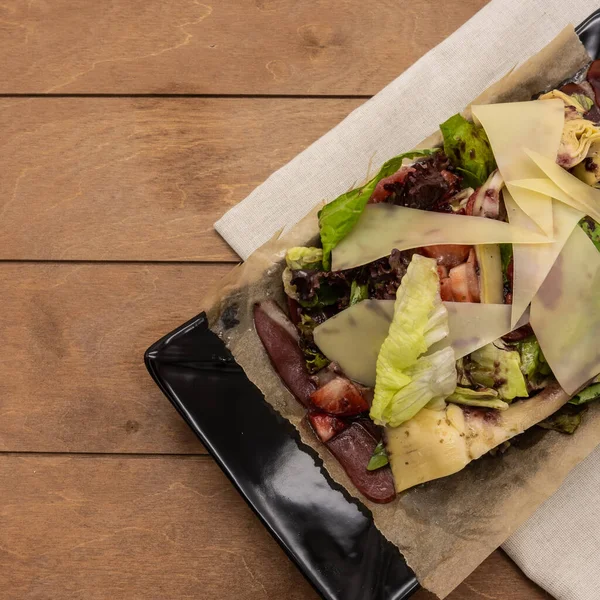 Smoked duck breast salad with cheese, green herbs and strawberries. Placed on a black rectangular plate. Background - brown boards, beige napkin. Top view