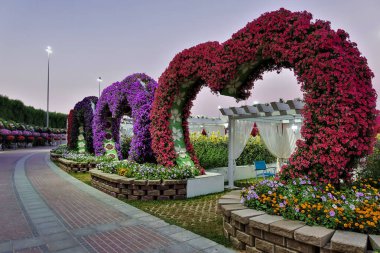 Park tasarımı. Taş patika bir virajın arkasında gizlidir. Yolun kenarında, canlı kırmızı ve morlardan yapılmış kalp şeklinde kemerler var. Derinliklerde perdeleri, sandalyeleri olan tenteler görülebilir..