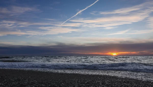 Deniz kenarında akşam alacakaranlığı. Mavi gökyüzünde sirus bulutları. Güneş suyun üzerinde alçalır. Plaj taşlarında köpük dalgaları. Karadeniz.