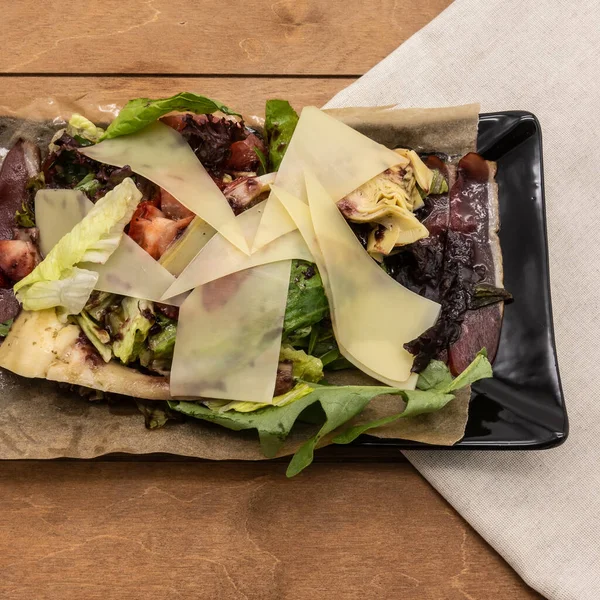 On a black rectangular plate, a salad of smoked duck breast, thin slices of cheese, fresh green herbs, strawberries. The background is a beige napkin, brown boards.