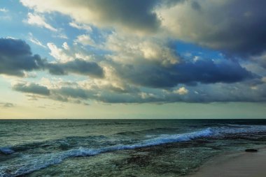 Karayip sahilinde bir sabah. Mavi gökyüzündeki kümülüs bulutları altınla işaretlenmiştir. Turkuaz dalgalar kumda köpürüyor. Meksika.
