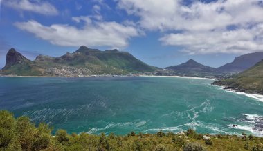 Cape Town 'daki körfez dağlarla çevrilidir. Sahilde plajı, kentsel alandaki evleri görebilirsiniz. Suyun turkuaz yüzeyinde beyaz köpük var. Gökyüzünde bulutlar var..
