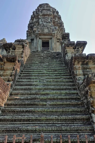 Weathered steps of a stone staircase lead to the top of the ancient temple. Tower with bas-reliefs and ornaments against the blue sky. The empty doorway is barred with bars. UNESCO. Angkor. Cambodia