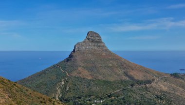 Aslan Kafası kayası, mavi gökyüzü ve Atlantik Okyanusu 'nun arka planında. Yamaçlarda yeşil bitki örtüsü. Virajlı yollar görülebilir. Cape Town 'da. Güney Afrika